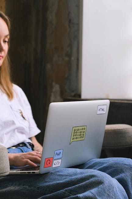 A young woman sits on a bench outdoors in a garden setting, partially visible in the frame. In front of her, a laptop rests on her lap, decorated with colorful stickers featuring coding language icons